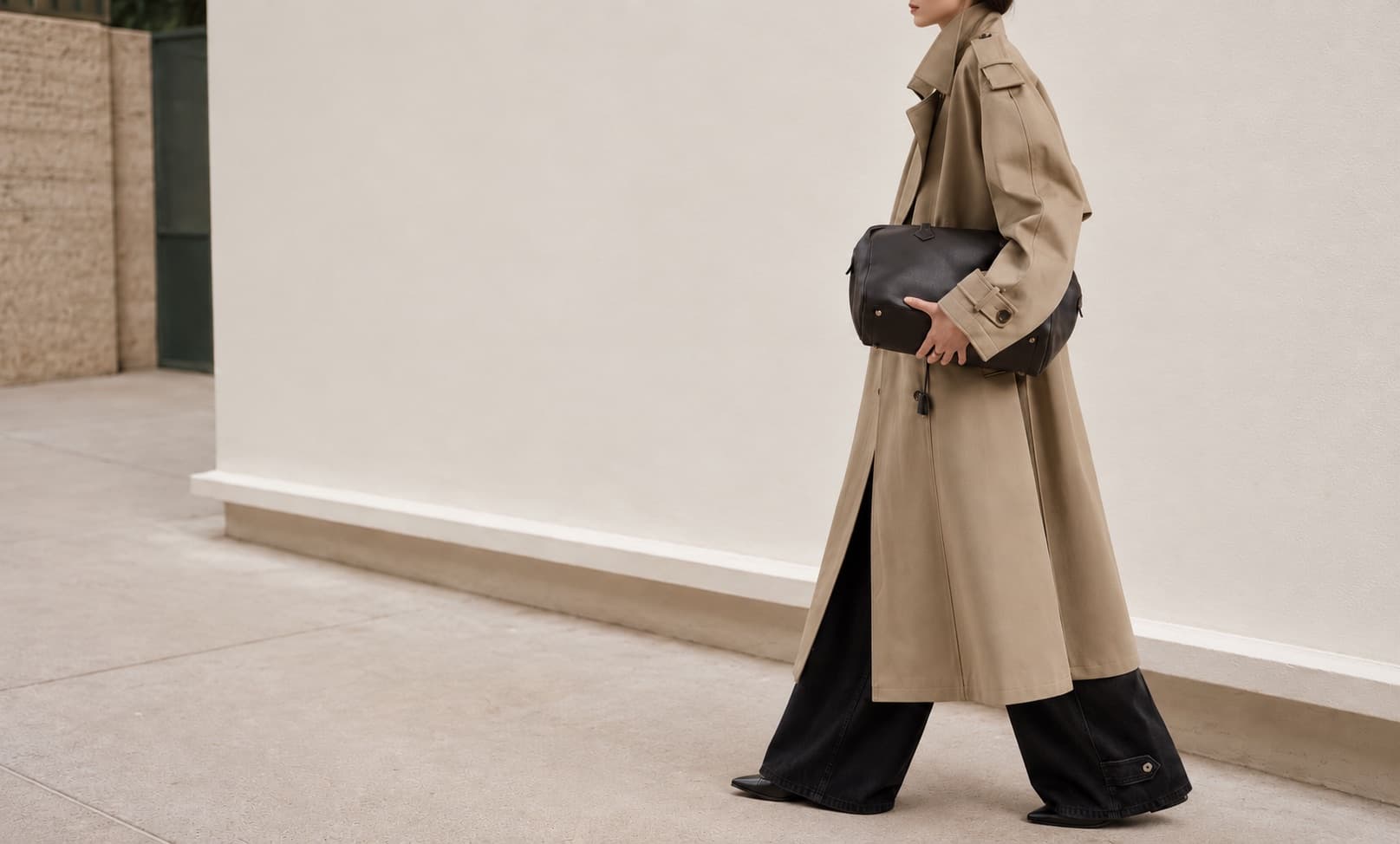 Model walking a sunlit wall in a trench coat, wide jeans and a leather bag